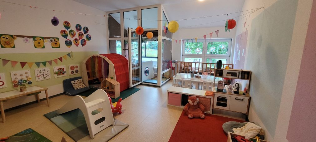 Blick in einen bunten Kindergartenraum mit Spielbereichen und Dekorationen.